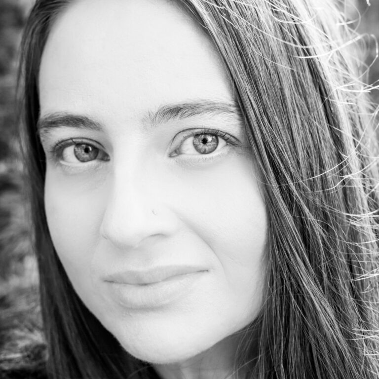 Portrait of a woman with long, straight hair, looking directly at the camera with a calm expression. The monochromatic image highlights the details of her face, including her light eyes and a small nose piercing. The background is softly blurred.