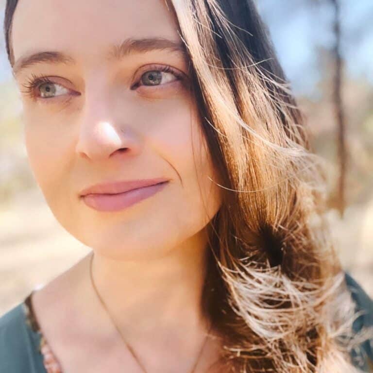 A woman with long brown hair and light-colored eyes looks into the distance with a serene expression. She is outdoors, bathed in soft sunlight, with a blurred background of trees. She wears a green top and a simple necklace.