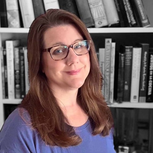 A woman with long brown hair and glasses smiles softly. She is wearing a blue top and stands in front of a bookshelf filled with books.