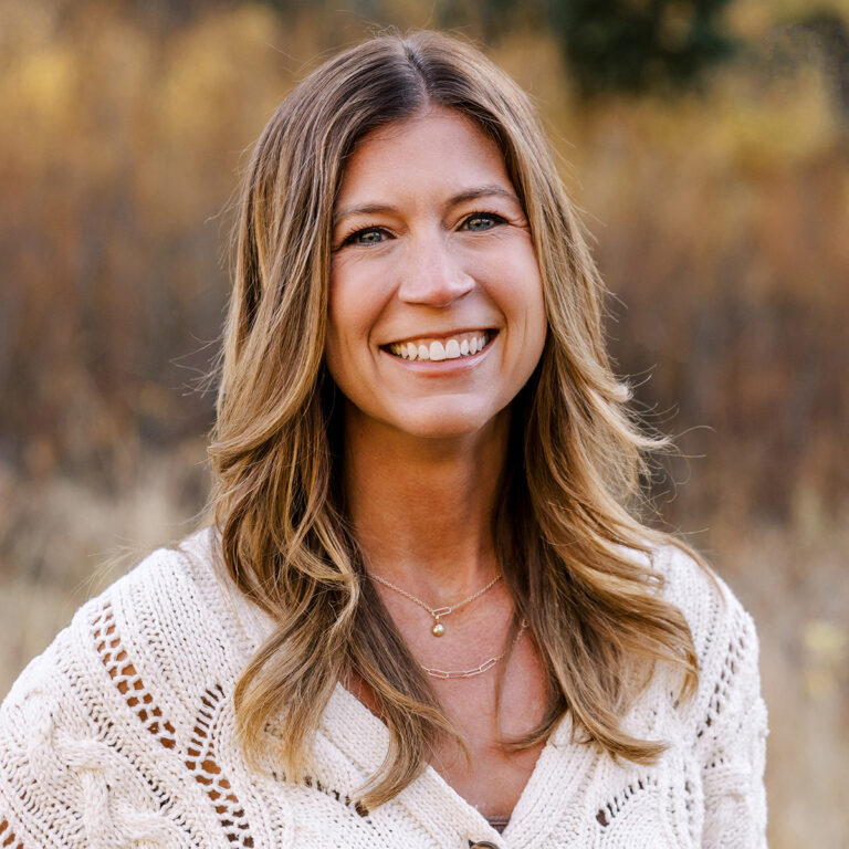 A woman with long, wavy brown hair smiles warmly at the camera. She is wearing a cream-colored, knit sweater and layered gold necklaces. The background features a blurred, natural setting with autumnal colors.