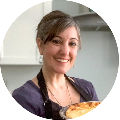 A woman smiling and wearing a black apron holds a freshly baked quiche or pie. She stands in a kitchen with white cabinets and a stove hood visible in the background.