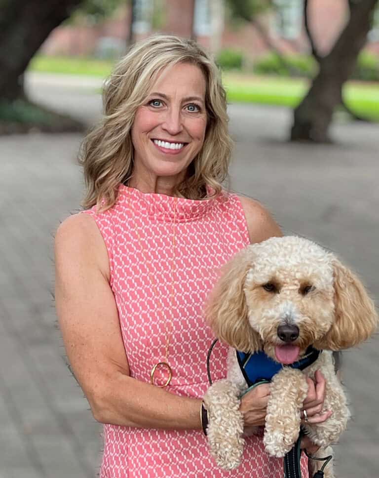 A woman with shoulder-length blonde hair, wearing a pink patterned sleeveless dress, stands outdoors holding a small, curly-haired dog in her arms. The dog is wearing a blue harness and has a happy expression. Trees and a paved walkway are in the background, setting the scene for some fun activities.
