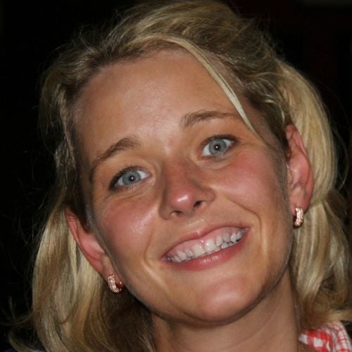 A person with blonde hair smiling at the camera. They are wearing earrings and a checkered shirt. The background is dark, highlighting their cheerful expression.