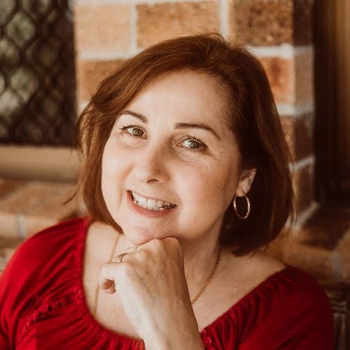 A person with shoulder-length brown hair is smiling, wearing a red top and gold hoop earrings. They are resting their chin on their hand. The background features a brick wall and a window with a metal grid.