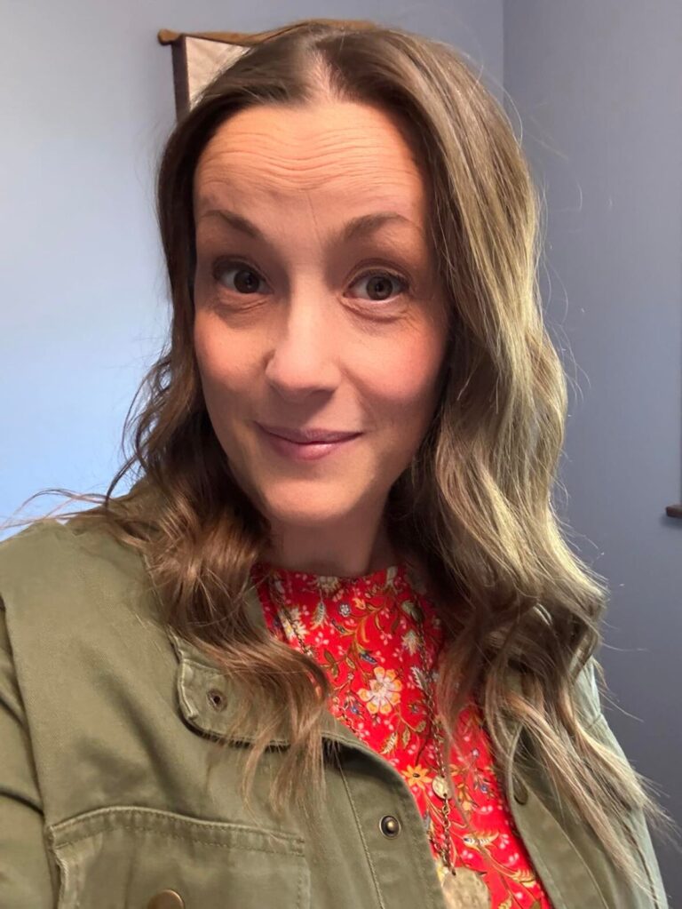 Woman with long wavy hair poses indoors, wearing a green jacket over a red floral top. She smiles slightly, looking at the camera. The background is a soft blue wall.