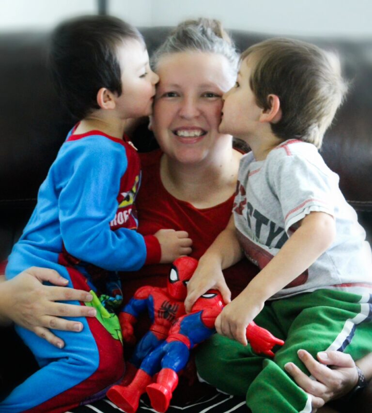A smiling woman sits on a couch with two young boys. The boys are each kissing her cheeks and holding a Spider-Man toy. One boy wears colorful pajamas, and the other wears a grey T-shirt with green pants. The scene is warm and cheerful.
