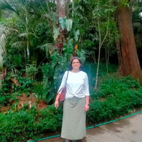 A person stands on a sidewalk in front of lush green foliage and tall trees, wearing a white top, long gray skirt, and carrying a brown bag. The background is a dense, leafy garden.