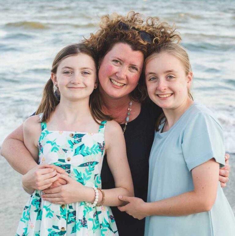 A woman with curly hair stands on a beach, smiling and hugging two young girls. The girl on the left wears a floral dress, while the girl on the right wears a light blue shirt. The ocean waves are visible in the background.