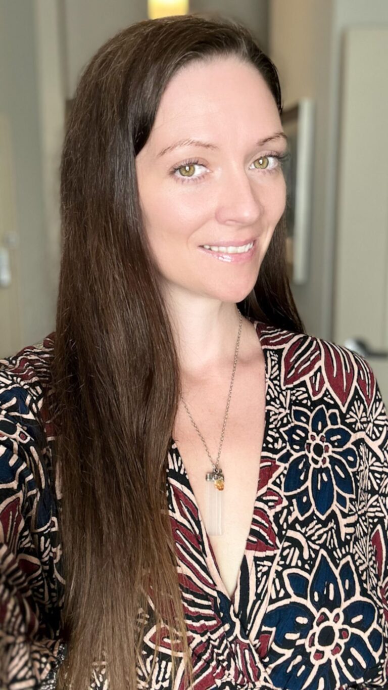 A woman with long brown hair smiles at the camera. She is wearing a patterned top with leaf and floral designs, and a necklace with a pendant. The background is blurred, focusing attention on her.