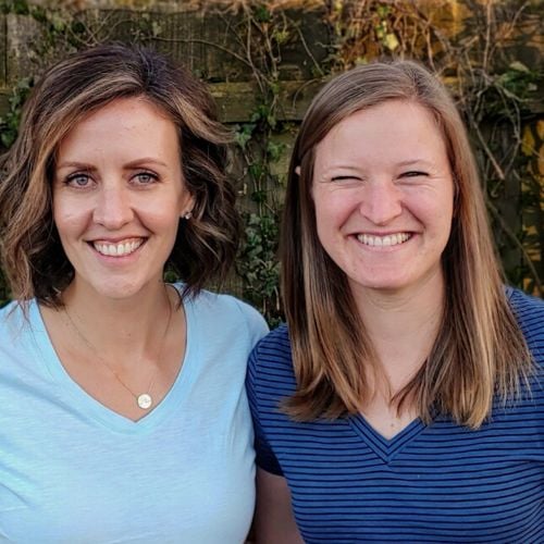 Two women smiling at the camera. The woman on the left has curly brown hair and is wearing a light blue top. The woman on the right has straight blonde hair and is wearing a dark blue striped top. They stand in front of a wooden fence with vines.