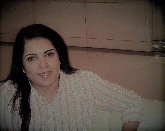 A person with long dark hair is sitting indoors, wearing a pink and white striped shirt. The background features a tiled wall and floor. The lighting is soft and warm.