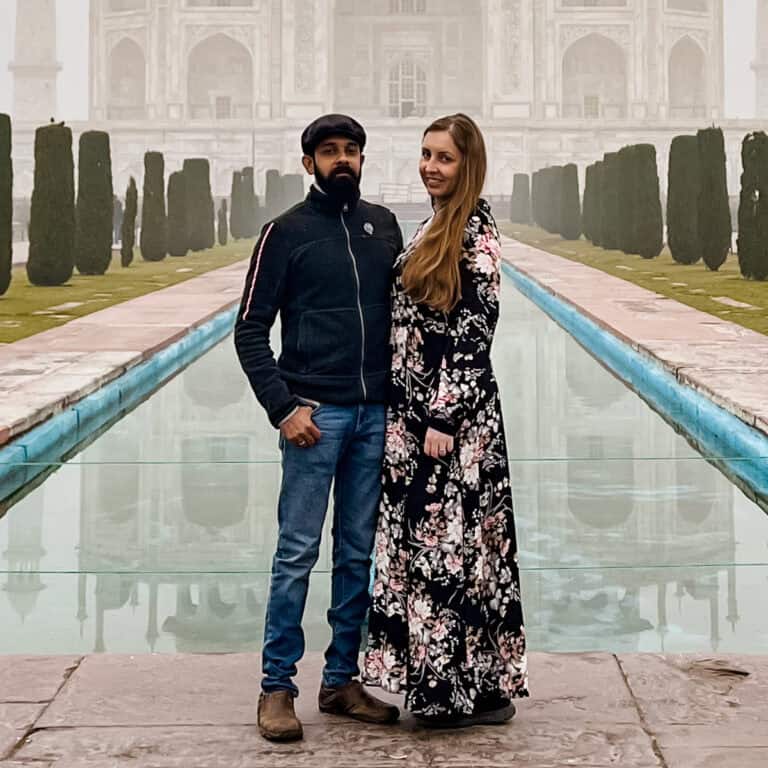 A couple stands in front of a long reflecting pool with the Taj Mahal in the background on a foggy day. The man is wearing a dark jacket, jeans, and a cap, while the woman is in a floral dress. They are both looking at the camera and smiling.