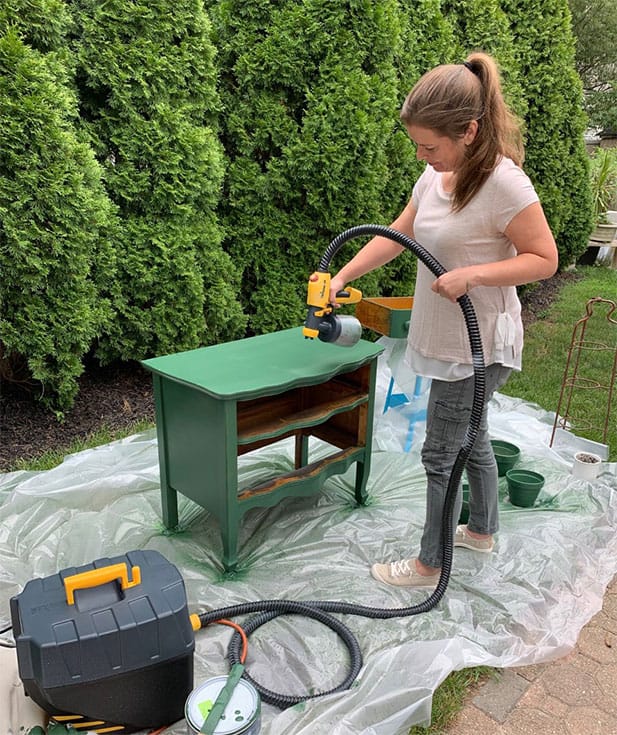 A woman paints a small wooden cabinet green using a paint sprayer. She is standing on a plastic sheet, with bushes in the background. A toolbox and paint can are on the ground nearby.