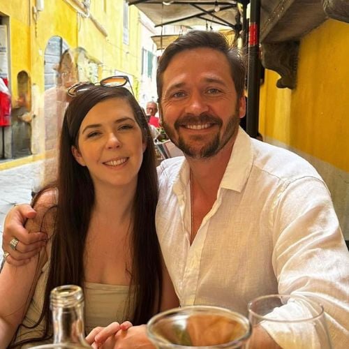 A smiling couple sits together at an outdoor café with yellow walls in the background. Both are wearing light-colored clothing. There are empty glasses and a bottle on the table in front of them.