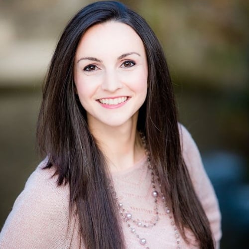 Smiling woman with long, straight dark hair, wearing a light pink sweater and a necklace, standing outdoors with a blurred background.