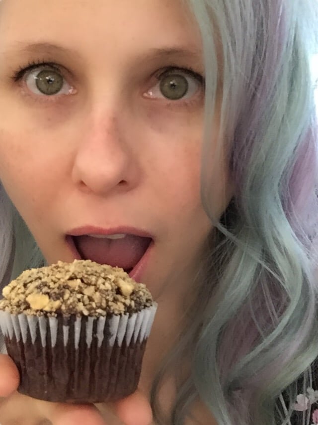 A person with long, wavy, pastel-colored hair holds a chocolate cupcake topped with crumbled topping close to their mouth, looking surprised or excited. The focus is on the cupcake and the person's facial expression.