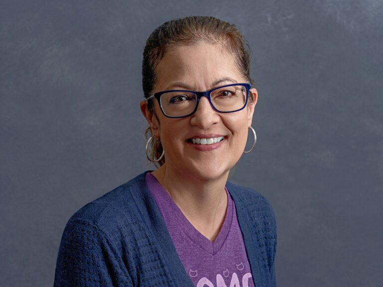 A person with glasses, wearing hoop earrings and a purple shirt with a dark blue cardigan, is smiling against a gray background.