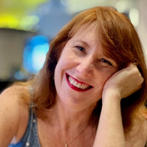 A smiling woman with red hair rests her head on her hand. She is wearing a sleeveless top and a necklace. The background is softly blurred with bright lights, creating a warm ambiance.