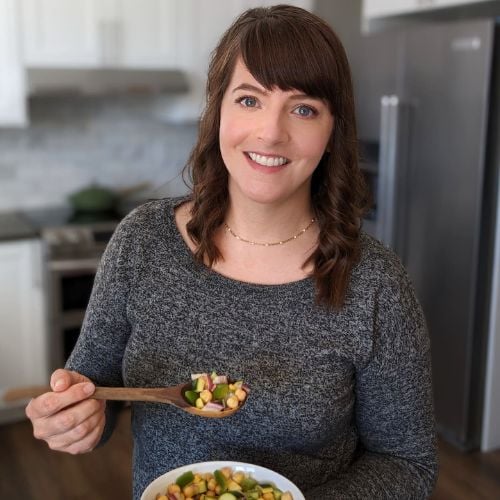 Person with brown hair in a gray sweater smiles while holding a bowl and spoon filled with a colorful salad, standing in a modern kitchen.