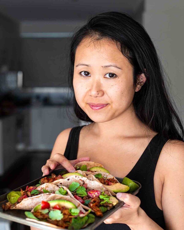 A person with long hair and a black tank top holds a plate of tacos filled with various colorful ingredients, including greens, meat, and lime wedges, in a brightly lit kitchen.