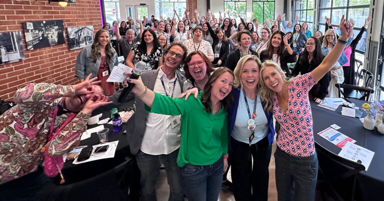 A group of excited people posing for a selfie indoors. They are smiling, cheering, and appear to be at a professional event or conference. Some hold up peace signs or wave, and there are tables with papers and drinks in the background.