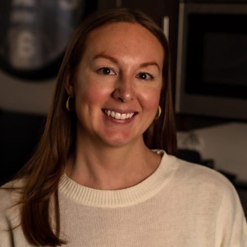 A person with long, straight hair, smiling and wearing a light-colored sweater and small gold hoop earrings. The background appears to be an indoor setting with dim lighting, featuring a blurred out clock and some dark furniture.