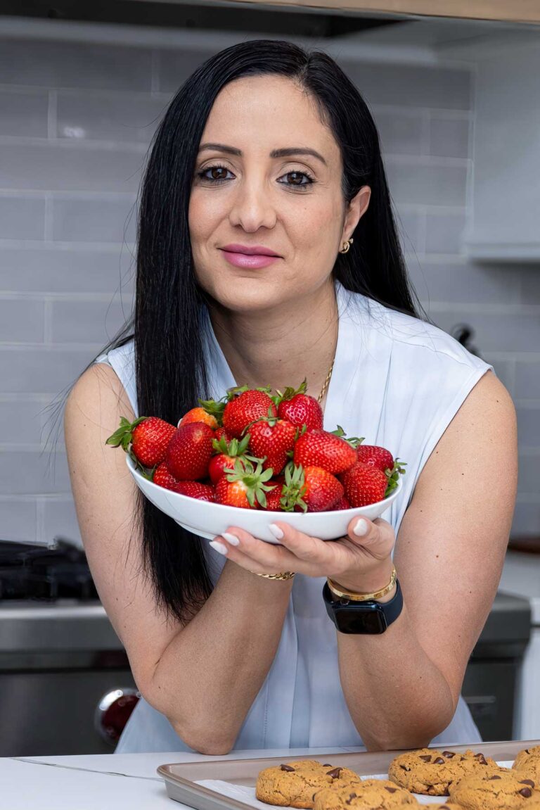 A woman holds a bowl of fresh strawberries, smiling gently. She is standing in a kitchen with a tray of cookies in front of her. The kitchen has a modern design with a tiled backsplash.