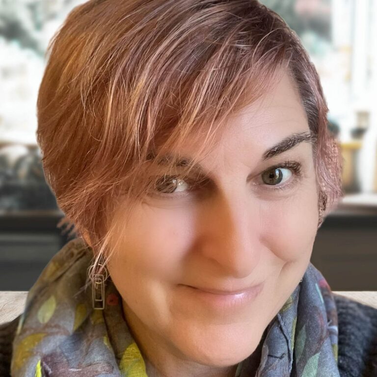 A person with short, light brown hair smiles softly at the camera. They are wearing a multicolored scarf and dangling earrings. The background is softly blurred.