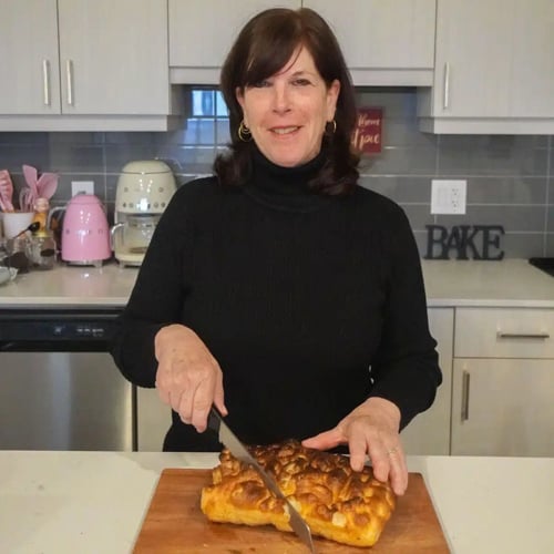 A woman with brown hair and a black turtleneck smiles as she slices bread on a wooden cutting board in a kitchen. The kitchen has a gray backsplash, and various kitchen items are visible in the background.