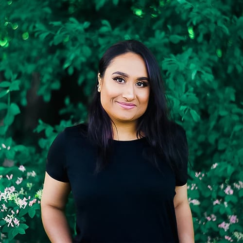 A person with long dark hair, wearing a black shirt, stands in front of lush green foliage. They are smiling and have their left hand resting by their side.