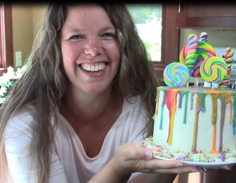 A woman smiles while holding a colorful cake decorated with rainbow lollipops and drips. The cake is white, with candy sprinkles at its base. She stands indoors against a kitchen background.