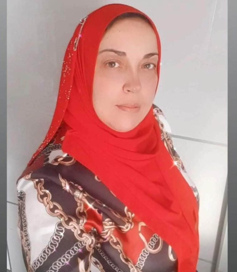 A woman wearing a red hijab and a patterned shirt with chain designs looks at the camera with a neutral expression, standing in front of a light-colored background.