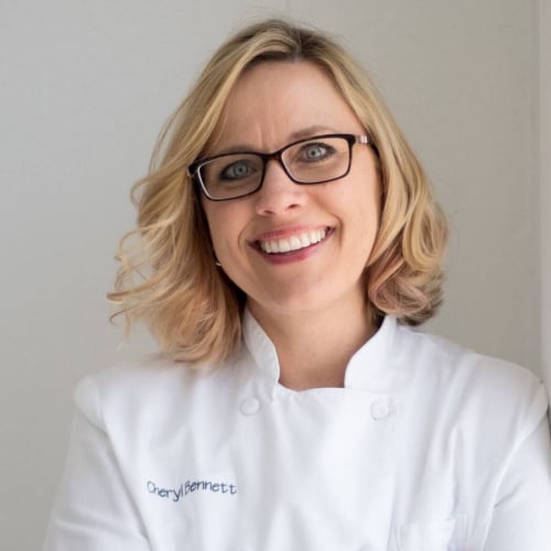A smiling woman with blonde hair and glasses is wearing a white chef coat embroidered with the name "Cheryl Bennett." She stands against a light-colored background.