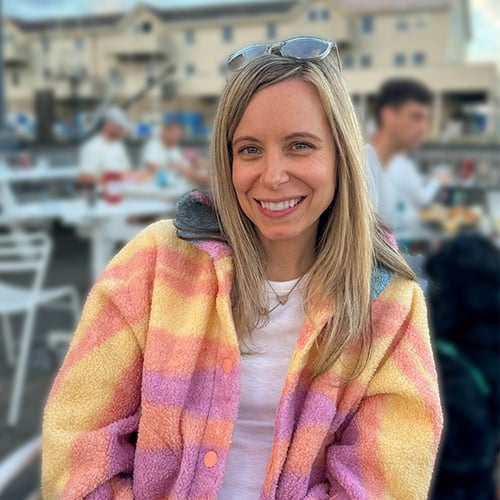 Smiling woman with long blonde hair, wearing a colorful striped jacket and sunglasses on her head, sits outdoors at a café. The background shows blurred people and buildings.
