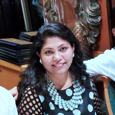 A woman with long, dark, wavy hair is sitting indoors, smiling at the camera. She is wearing a black and white polka dot top and a large, ornate necklace. There is intricate wood carving visible in the background.