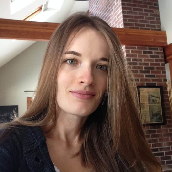 A person with long brown hair and a slight smile is indoors. They wear a dark sweater, and there's a brick wall and framed pictures in the background. Natural light from a window above lights up the space.