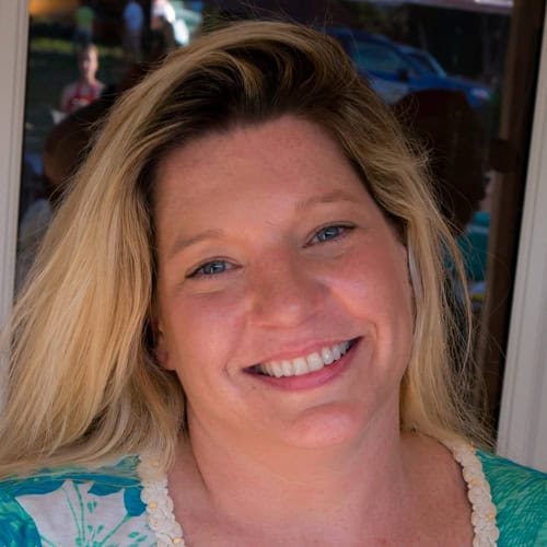 A woman with long blonde hair is smiling at the camera. She is wearing a green and white top. The background is slightly blurred but appears to be outdoors with a few other people in the distance.