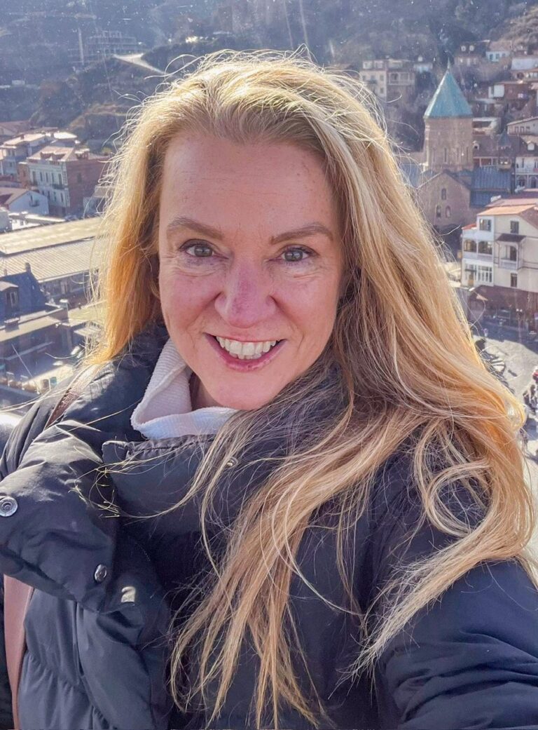 A person with long blonde hair smiles at the camera, wearing a black puffer jacket. The background features a scenic view with historic buildings and a church with a dome, under a clear sky.