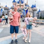 A family of four stands in front of a castle at a theme park. The father holds a child, and the mother holds another child. The young girl between them holds both parents' hands. The sky is overcast, and the castle is adorned with blue and white accents.