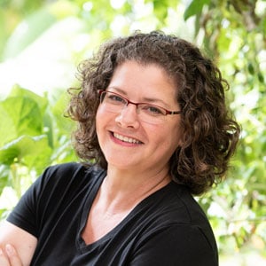 Smiling person with curly brown hair and glasses, wearing a black shirt, standing against a leafy green background.