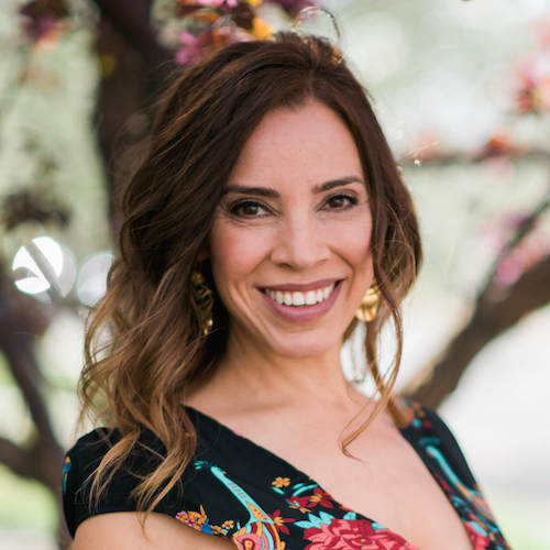 Smiling woman with long brown hair wearing a floral dress stands in front of a blurred background with tree branches and pink blossoms.